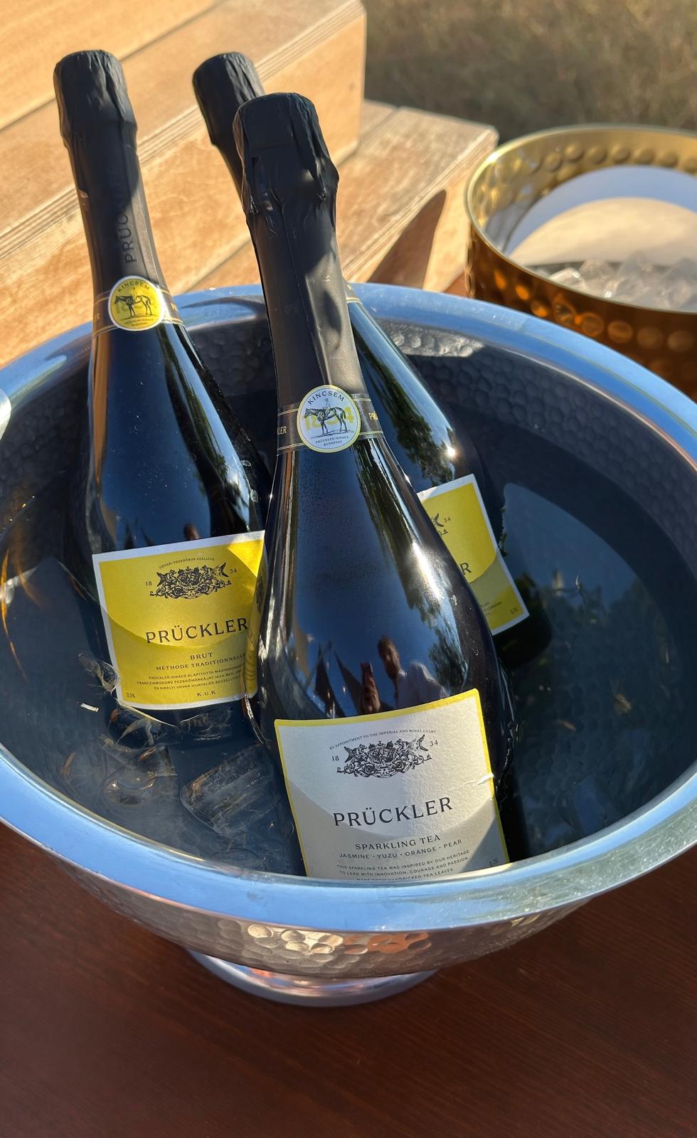 Three Prückler sparkling wine bottles in a hammered metal ice bucket on a wooden surface, with warm natural sunlight casting shadows.